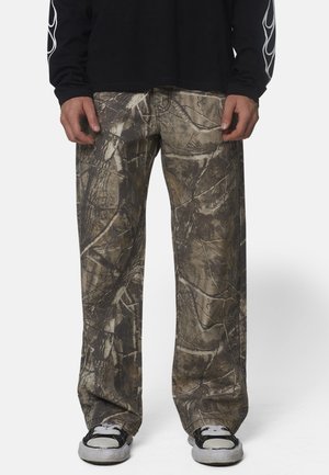 Homme portant un pantalon camouflage marron et vert à jambes larges, des baskets noires et blanches et un t-shirt noir à manches longues avec un motif blanc sur les manches.