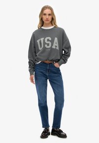 Grijze sweatshirt met "USA" in crème gedrukt, gecombineerd met een jeans in blauw. Zwarte en bruine schoenen passen goed bij de outfit. Eenvoudig, casual ontwerp.