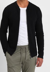 Černá bomber bunda na zip z texturované pleteniny, s kulatým výstřihem a přiléhavými manžetami, v kombinaci s bílým tričkem a světlými kalhotami.