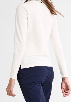 Femme de dos, portant un haut blanc à manches longues et un pantalon bleu foncé, debout devant un fond clair et uni.