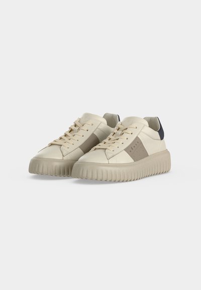 Paire de baskets basses beiges avec semelles épaisses striées, panneaux latéraux taupe, lacets beiges et accents noirs au talon sur un fond clair.