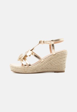 Sandale compensée avec des brides en or métallisé, une boucle à la cheville, des décorations florales sur le dessus et une semelle plateforme en jute tressé.