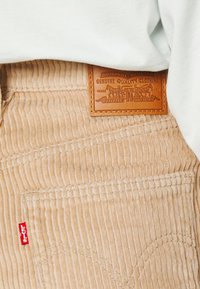 Pantalons en velours côtelé beige avec des côtes verticales, dotés d'un label en cuir brun et d'une étiquette rouge Levi's sur le côté, partiellement cachés par un haut clair.