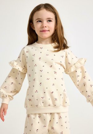 Ragazza giovane con capelli castani lunghi che indossa una felpa color crema e pantaloni con piccole stampe floreali e dettagli arricciati sulle maniche.