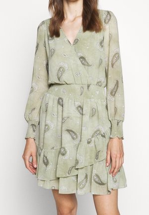 Kobieta ubrana w jasnozieloną sukienkę z długim rękawem z motywem paisley, z marszczoną talią i warstwową falbaną, stojąca na jednolitym tle.