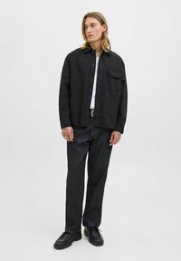 Young man with long hair wearing a black button-up shirt, white undershirt, dark jeans, black shoes, and standing against a plain white background.