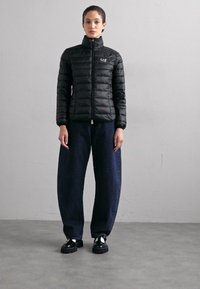 Črna pufastajna jakna z visokim ovratnikom in sprednjim zadrgo, kombinirana s temno modrimi širokimi jeans hlačami in debelimi črnimi čevlji. Gladka, prešita tekstura.
