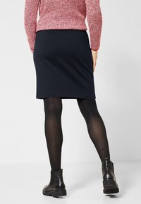 Tmavě modrá sukně z měkké tkaniny, s úzkým střihem a délkou ke kolenům. Nošena s černými punčocháči a kotníkovými botami, zdůrazňující čisté linie.