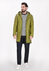 Zielona wodoodporna kurtka z kapturem, z zatrzaskami i kieszeniami, noszona na czarno-białą w paski koszulę i ciemne dżinsy.