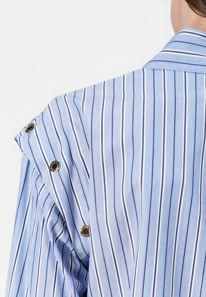 Blauw gestreept shirt met verticale lijnen, voorzien van een brede kraag en decoratieve knopen op de schouder. Het materiaal lijkt gestructureerd en licht van gewicht.