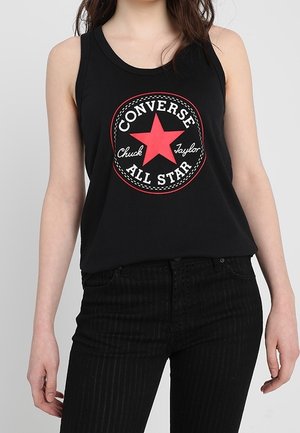 Vrouw die een zwart mouwloos topje draagt met een rood Converse All Star-logo en zwarte hoge taille broek tegen een effen achtergrond.