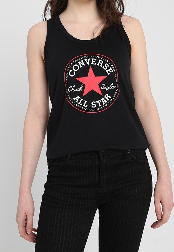Femme portant un haut noir sans manches avec le logo rouge Converse All Star et un pantalon noir taille haute sur un fond uni.