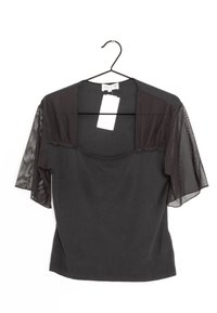 Svart blus med korta genomskinliga nätärmar, fyrkantig halsringning och en kropp av enfärgad tyg. Hänger från en svart krok med en storlekslapp fäst.