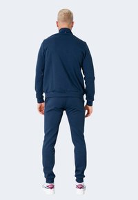Chándal deportivo azul marino con chaqueta con cremallera y pantalones ajustados. Presenta una textura suave y puños acanalados, combinado con zapatillas blancas.