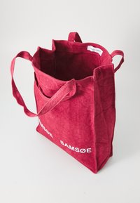 Borsa a tracolla rossa in tessuto con due spallacci, apertura superiore e tasca frontale. Testo bianco "SAMSOE SAMSOE" sul lato. Texture liscia.