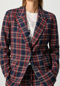 Blazer a quadri rossi e blu con una vestibilità sartoriale, dotato di rever a punta, due bottoni e una texture del tessuto liscia. Indossato sopra una maglietta grafica.