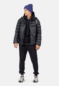 Sort pufferjakke, navy hættetrøje, sorte sweatpants og grå sneakers. Blå ribbet beanie. Rygsæk med skulderremme.