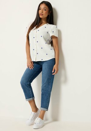 Frau trägt eine weiße kurzärmelige Bluse mit blauen Blumen, hochgekrempelte blaue Jeans und weiße Sneakers, steht vor einer weißen Wand.