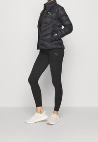 Czarna kurtka puchowa z wysokim kołnierzem, pikowany wzór; czarne legginsy z dyskretnym logo; jasne trampki z siateczki, różowe akcenty.