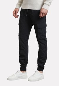 Pantalones cargo negros hechos de una tela liviana, con múltiples bolsillos, puños elásticos y una cinturilla ajustable con cordón. Llevados con zapatillas blancas.