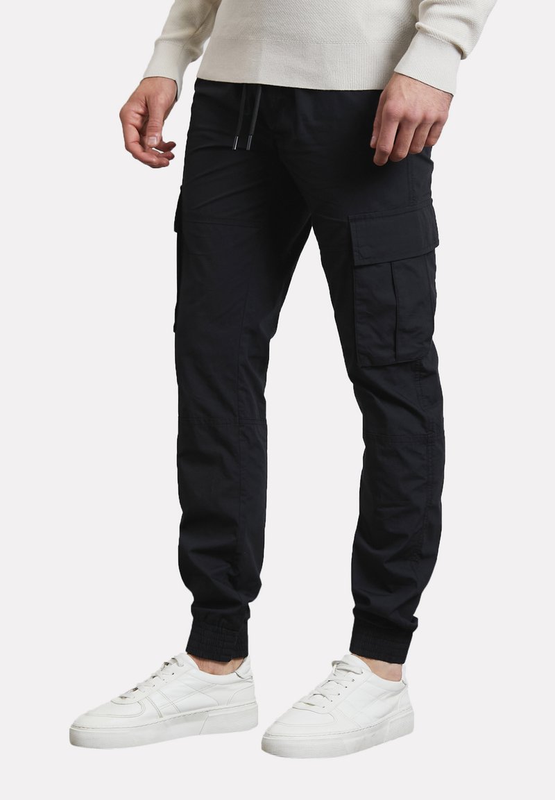 Pantalones cargo negros hechos de una tela liviana, con múltiples bolsillos, puños elásticos y una cinturilla ajustable con cordón. Llevados con zapatillas blancas.
