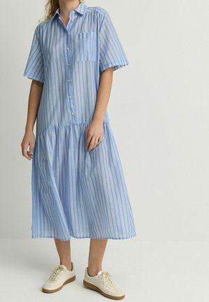 Frau trägt ein knielanges Kleid mit hellblauen und weißen vertikalen Streifen, kurzen Ärmeln und weißen Turnschuhen vor einfarbigem Hintergrund.