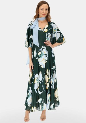 Kobieta ubrana w ciemnozieloną suknię maxi z dużymi białymi i żółtymi kwiatowymi wzorami oraz jasnoniebieską apaszką na szyi, pozująca na tle jednolitego tła.