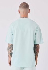 Camiseta de manga corta verde menta en un corte holgado. Tejido suave, cuello redondo, con tatuajes visibles en el brazo. Diseño sencillo, sin patrones ni acentos.