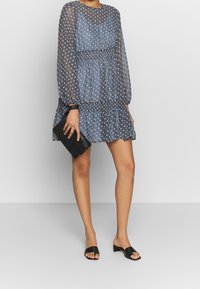 Blå mönstrad klänning med genomskinliga långa ärmar, smockad midja och volangkant. Matchad med en svart kuvertväska och platta svarta sandaler.