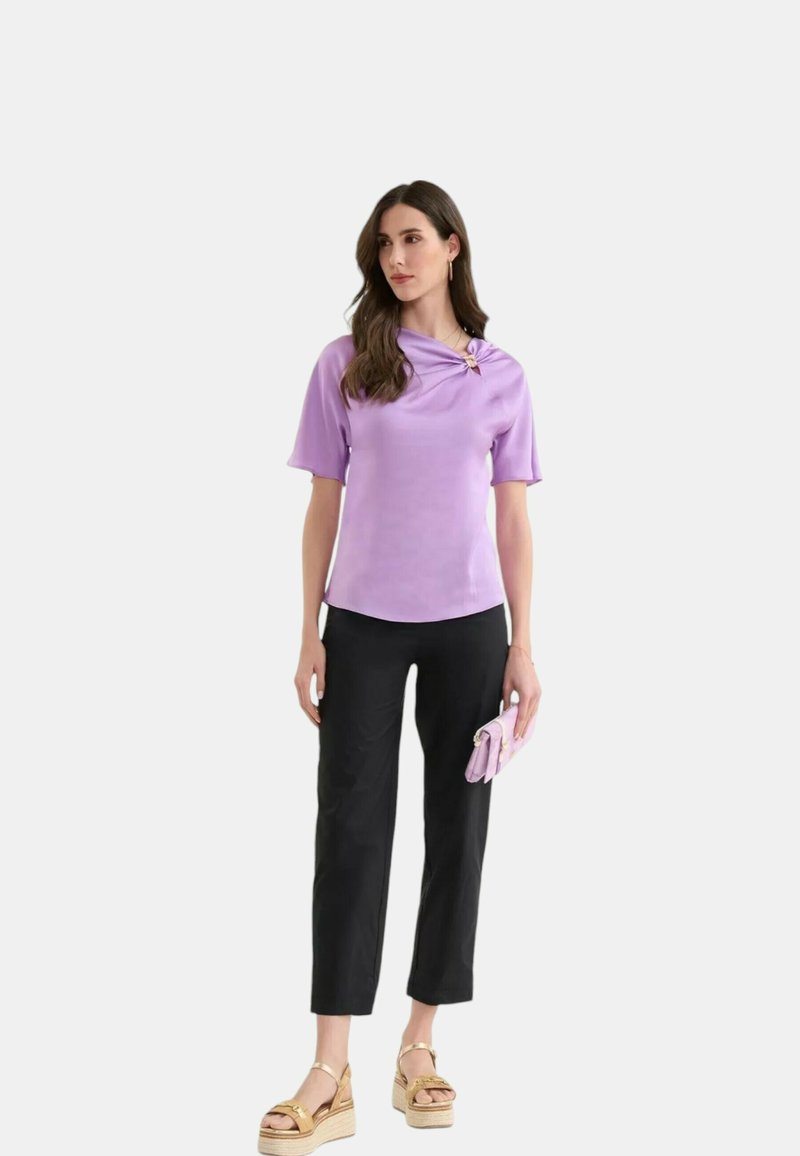 Top lavanda a maniche corte con scollo drappeggiato e dettaglio nodo laterale, abbinato a pantaloni neri a 3/4 e sandali con zeppa beige.