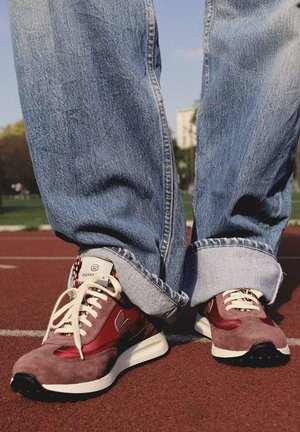 Personne portant un jean bleu retroussé et des baskets en daim rouge debout sur une piste de course extérieure.