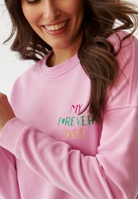 Světle růžová mikina s vyšitým textem "MY FOREVER DATE" v červené, zelené a zlaté barvě. Volný střih s žebrovanými manžetami a kulatým výstřihem.
