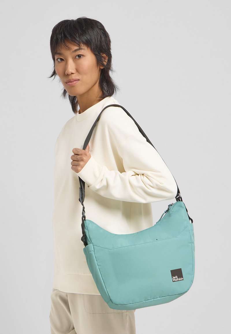 Eine türkisfarbene Schultertasche aus weichem Stoff mit einem geschwungenen Design, ausgestattet mit einem schwarzen Tragegurt und einem Jack Wolfskin Logo.