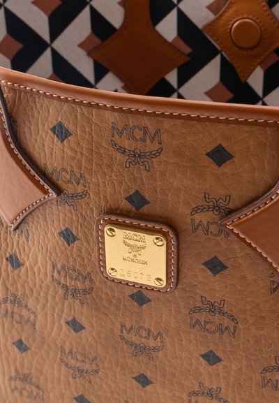 Καφέ δερμάτινη τσάντα MCM με επαναλαμβανόμενο μαύρο μοτίβο λογότυπου και χρυσή μεταλλική πλακέτα με την επιγραφή "MCM München" και σειριακό αριθμό.