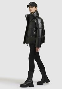 Zwarte pufferjack met hoge kraag, glanzende afwerking en zijzakken, gecombineerd met zwarte leggins en veterboots.