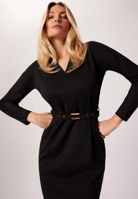 Czarna sukienka z długimi rękawami, dekoltem w kształcie litery V, z talią ściągniętą paskiem z złotymi akcentami. Gładki materiał z elegancką, dopasowaną sylwetką.