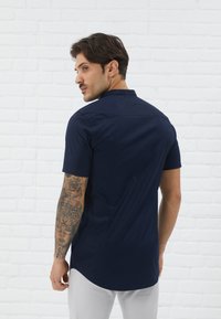 Navy short-sleeve shirt made of smooth fabric, featuring a collar and back yoke design. Model shows tattoos on arms against a white brick wall.