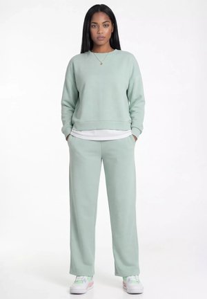 Młoda kobieta ubrana w jasnozieloną bluzę i pasujące szerokie spodnie z białymi trampkami, stoi z rękami w kieszeniach na białym tle.