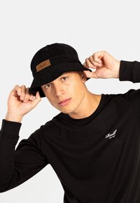 Sombrero tipo bucket negro con una tela texturizada y un parche de cuero marrón que muestra el nombre de la marca. El modelo lleva una camiseta de manga larga negra.