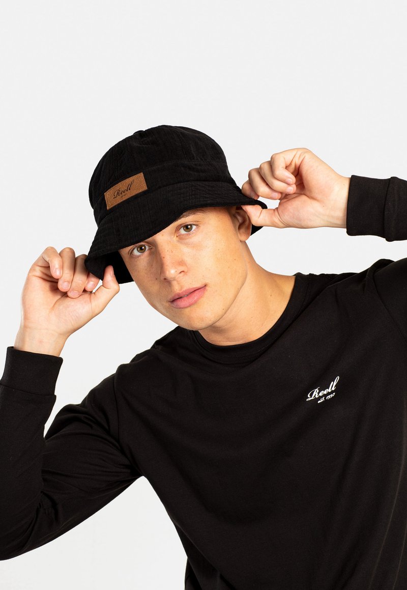 Sombrero tipo bucket negro con una tela texturizada y un parche de cuero marrón que muestra el nombre de la marca. El modelo lleva una camiseta de manga larga negra.