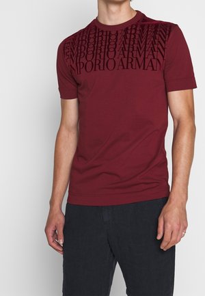 Mężczyzna ubrany w bordową koszulkę z krótkim rękawem z czarnym wzorem napisu "EMPORIO ARMANI" na klatce piersiowej oraz czarne spodnie.