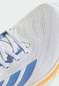 Primer plano de una zapatilla de running blanca con rayas azules, cordones blancos y detalle de suela naranja que muestra una textura de malla y agujeros de ventilación.