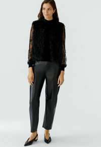 Schwarze Kunstfellweste über einem schwarzen Langarm-Oberteil, kombiniert mit formschlüssigen, schwarzen Kunstlederhosen und schwarzen Slingback-Highheels mit spitzer Zehenpartie.