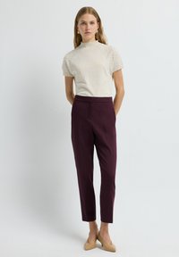 Vrouw die tegen een witte achtergrond staat, gekleed in een crèmekleurig getextureerd T-shirt met korte mouwen, donkerpaarse op maat gemaakte broek en beige hakken.