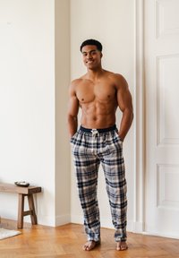 Kaal torso man staat op blote voeten in een geruite pyjamabroek, glimlachend met zijn handen in de zakken in een heldere kamer met een houten vloer en een bank.