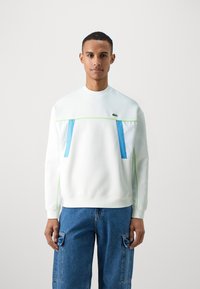 Sweat-shirt à col rond en tissu blanc avec des poches avant bleu et blanc, des accents vert fluo et des poignets côtelés. Porté avec un jean cargo bleu.