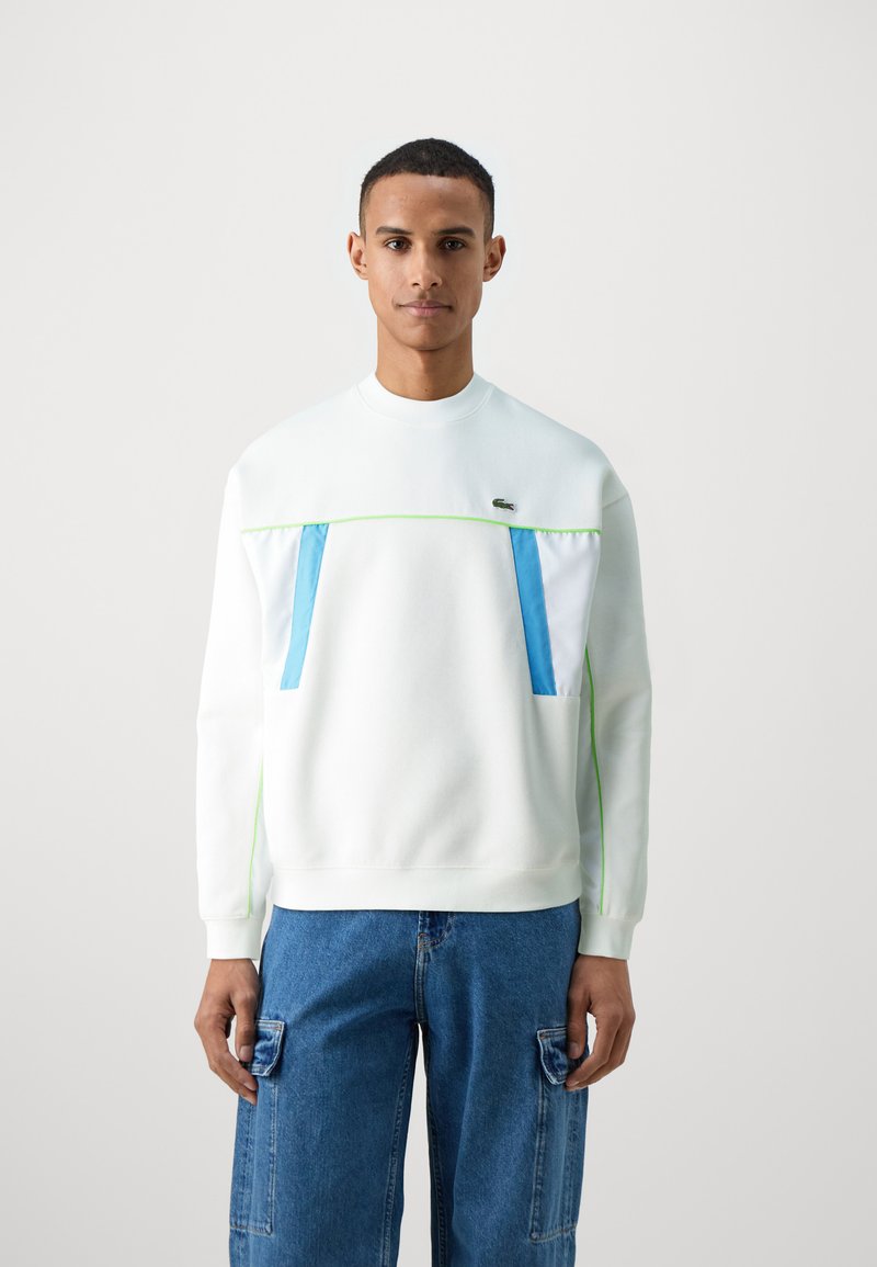 Sweat-shirt à col rond en tissu blanc avec des poches avant bleu et blanc, des accents vert fluo et des poignets côtelés. Porté avec un jean cargo bleu.