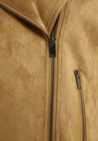 Beige mockaljacka med matt yta, med silverfärgade dragkedjor fram och i sidofickorna. Ren linje och vinkelriktig design.