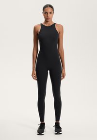 Fekete sportos bodyn, magas nyakkivágással, testhezálló szabással és sima textúrával. Fekete futócipővel viselve. Egyszerű és dísztelen stílus.