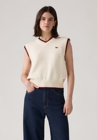 Suéter de punto sin mangas en color crema con escote en V, que presenta ribetes en rojo y azul marino, y un pequeño emblema de un perro marrón. Combinado con unos pantalones vaqueros de pierna ancha en azul oscuro.